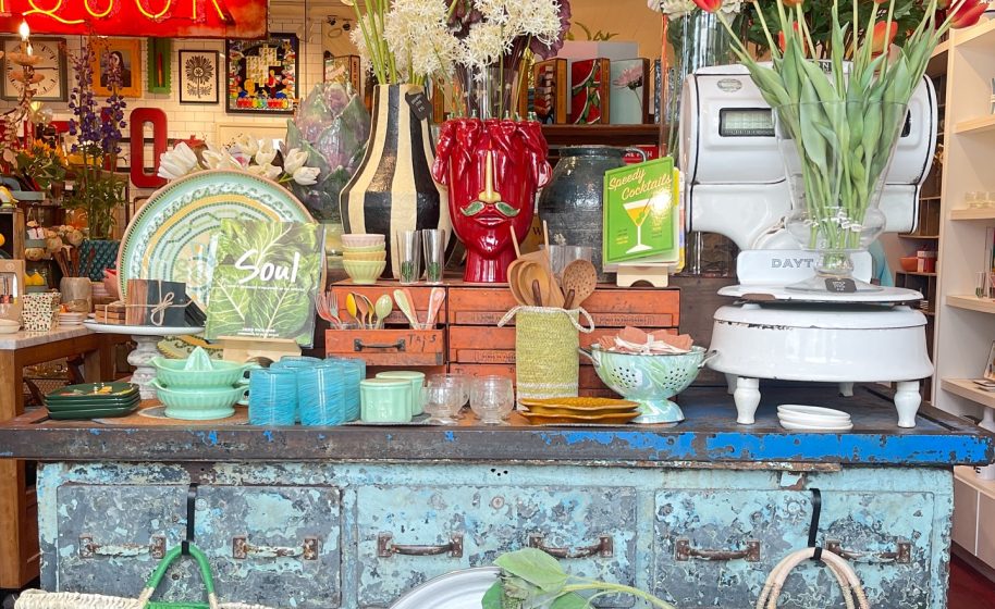 Colorful boutique display with flowers, ceramics, glassware, and vintage scale on distressed blue counter.