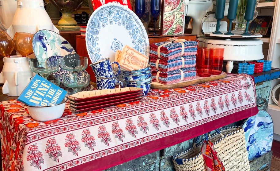 Colorful coastal gift shop display with patterned dishes, glassware, linens, and woven baskets on table.