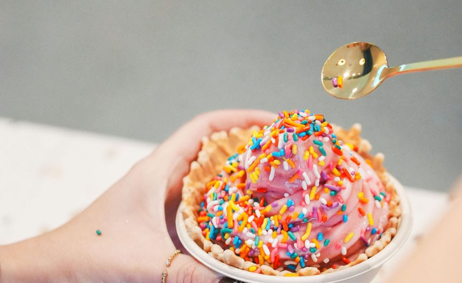 Pink frozen yogurt with rainbow sprinkles in waffle bowl, held in hand.