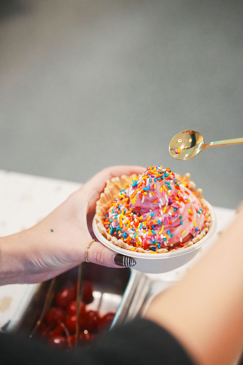 Pink frozen yogurt with rainbow sprinkles in waffle bowl, held in hand.