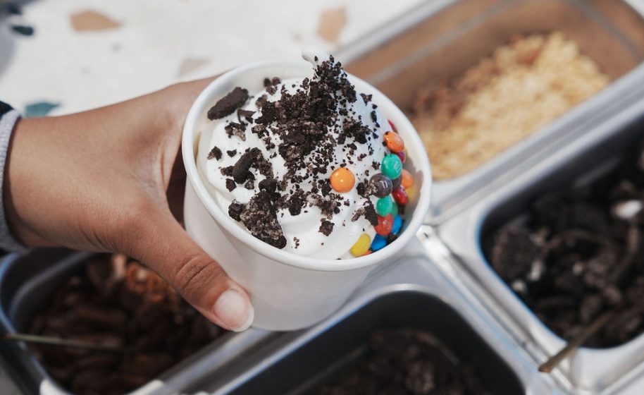 Hand holding frozen yogurt topped with crushed cookies and colorful candy pieces.