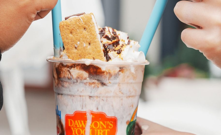 Hands hold a whipped-cream-topped frozen yogurt sundae with graham cracker and chocolate drizzle.