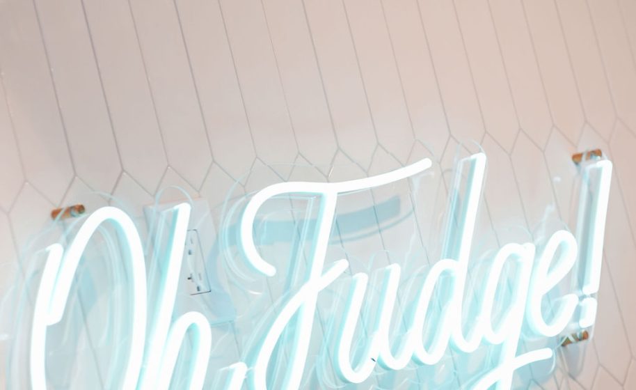 Neon script sign reading “Oh Fudge!” glows against a white tiled wall.