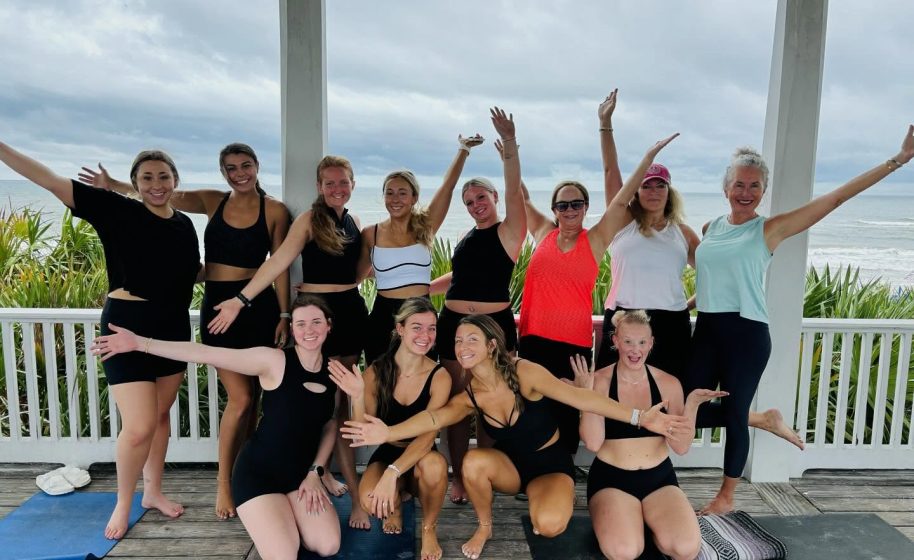 Group of women pose joyfully in athletic wear after outdoor yoga session on covered porch facing the ocean.
