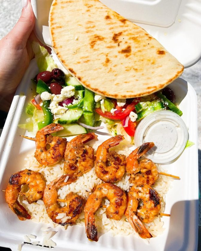 Grilled shrimp skewers served over white rice with Greek salad, pita bread, and a side dressing.