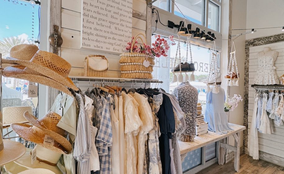 Straw hats, woven bags, and flowy dresses are displayed in a bright boutique with rustic decor and sunlight.