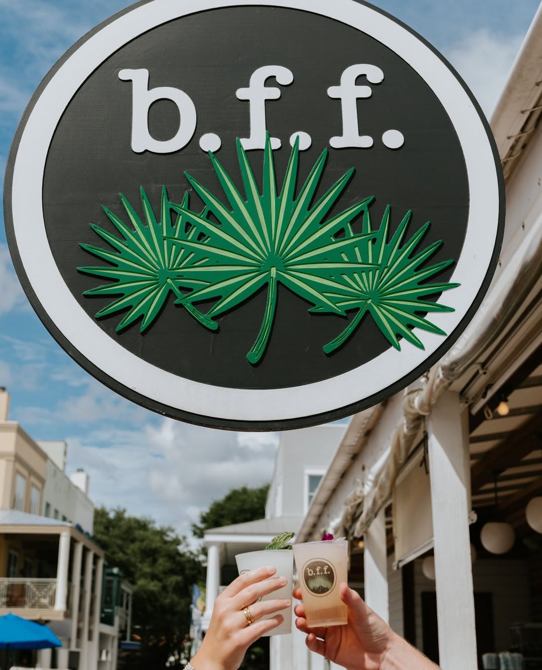 Large “b.f.f.” sign with green palm leaves above, hands holding branded drinks underneath, outdoor setting visible.