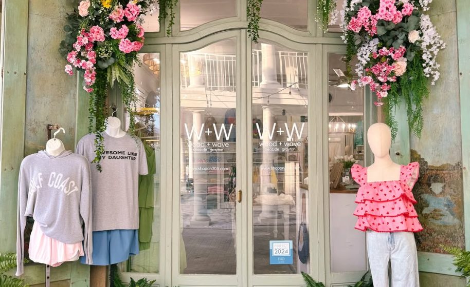 Boutique storefront with mannequins in casual outfits, mint doors, and hanging pink floral arrangements above.