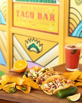 Colorful tacos and chips on wooden tray with orange half, jalapeño, and chilled cocktail beside Taco Bar sign.