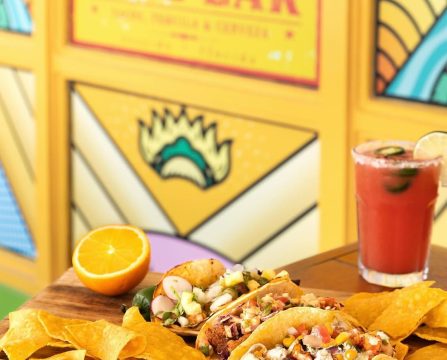 Colorful tacos and chips on wooden tray with orange half, jalapeño, and chilled cocktail beside Taco Bar sign.