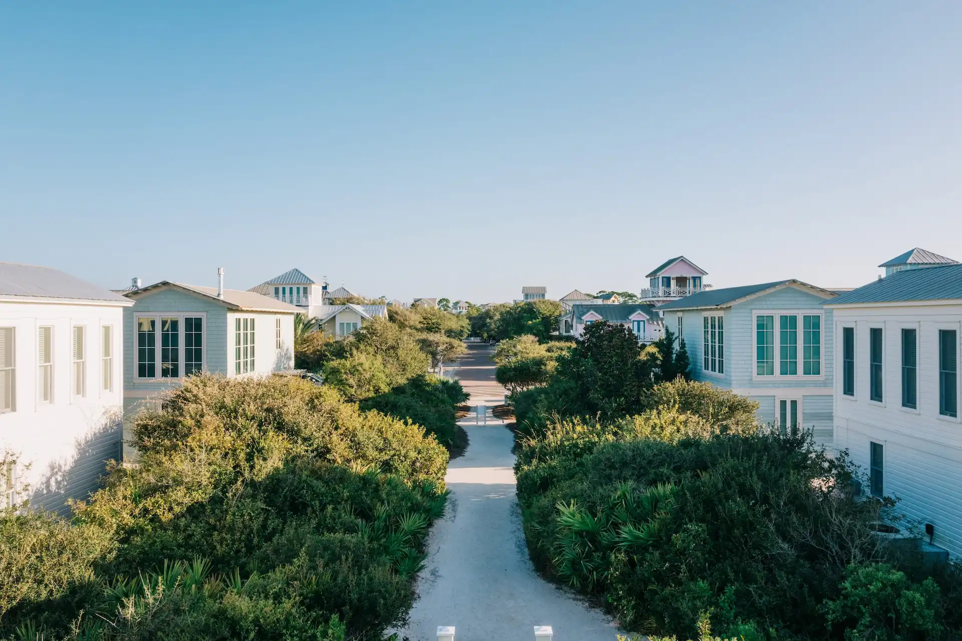 Pathway lined with greenery leading between pastel coastal houses under a clear blue sky near the beach.