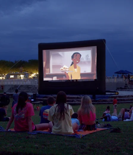 People sitting on grass at night watching animated movie outdoors on large inflatable screen in park setting.