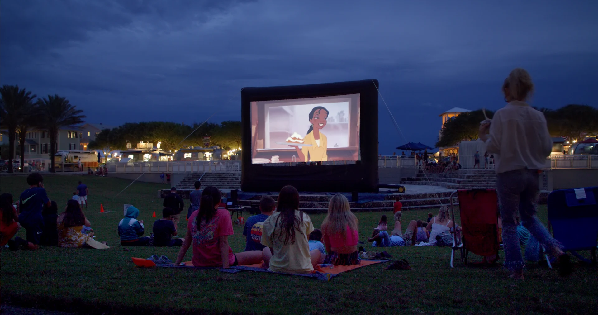 People sitting on grass at night watching animated movie outdoors on large inflatable screen in park setting.