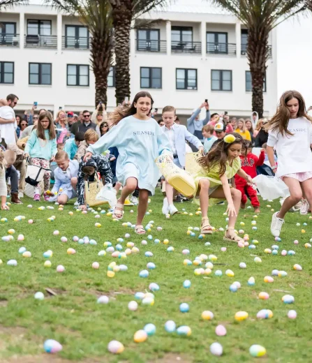 Children run across a grassy lawn collecting colorful Easter eggs as a crowd watches near palm trees and a resort building.