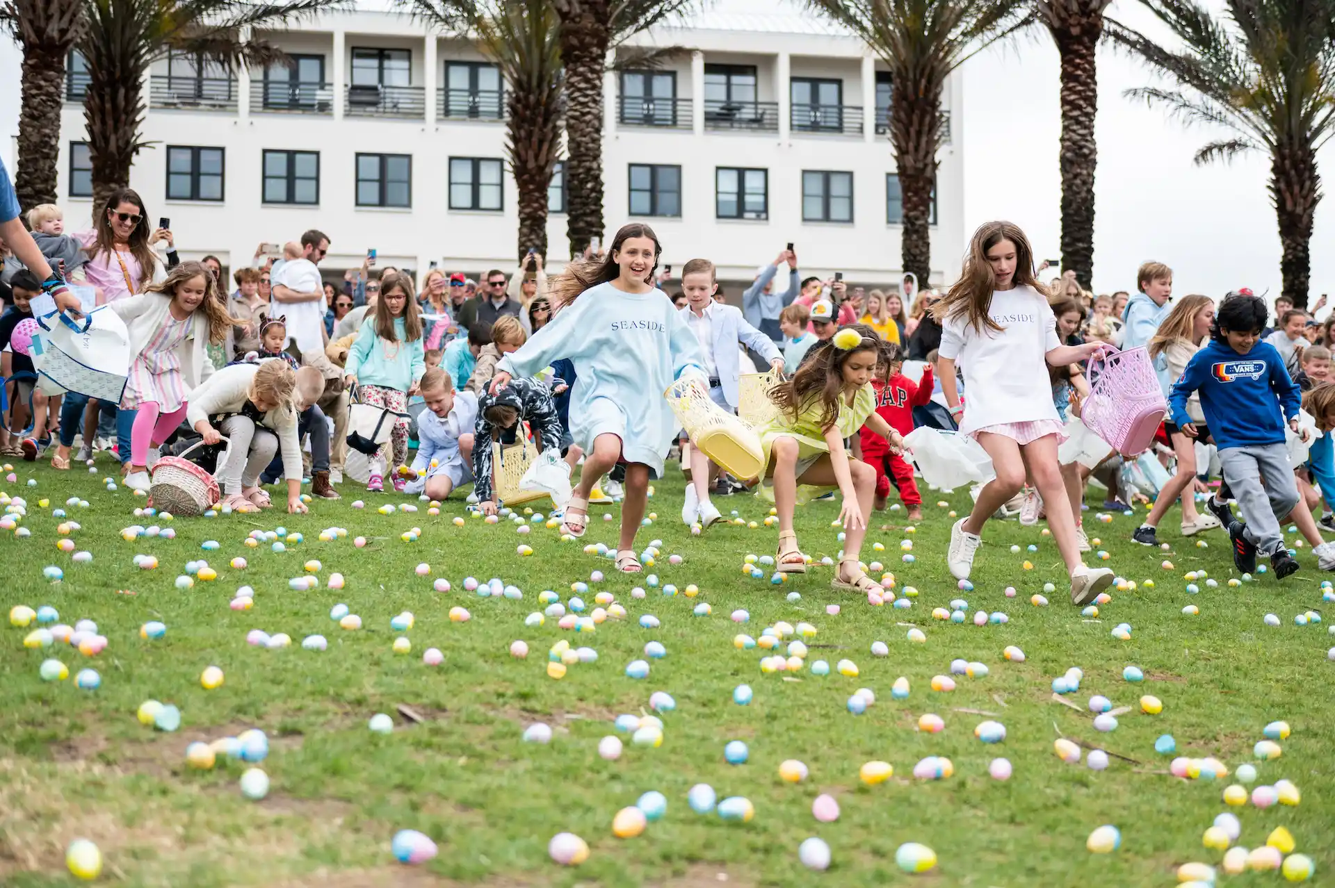 Children run across a grassy lawn collecting colorful Easter eggs as a crowd watches near palm trees and a resort building.