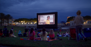 People sitting on grass at night watching animated movie outdoors on large inflatable screen in park setting.