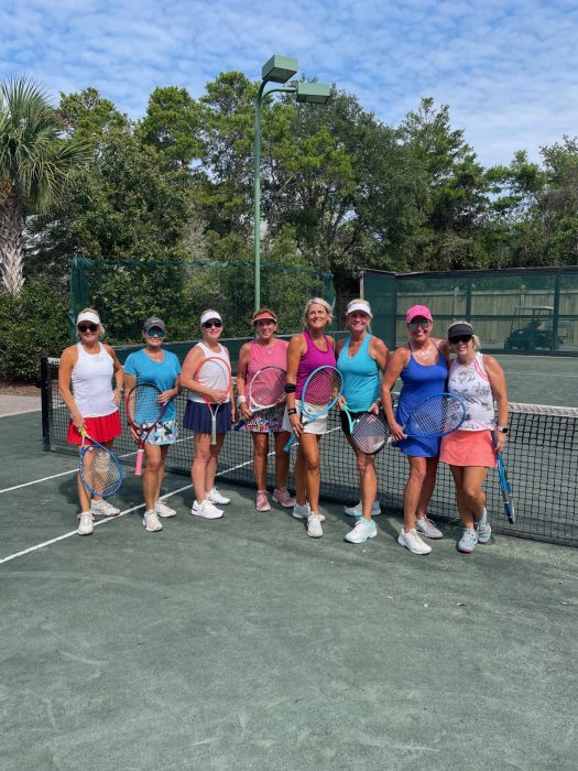 Group of women holding tennis rackets posing together on an outdoor tennis court under blue sky.