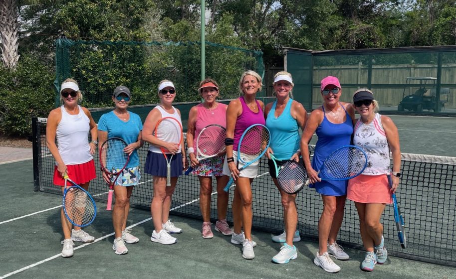 Group of women holding tennis rackets posing together on an outdoor tennis court under blue sky.