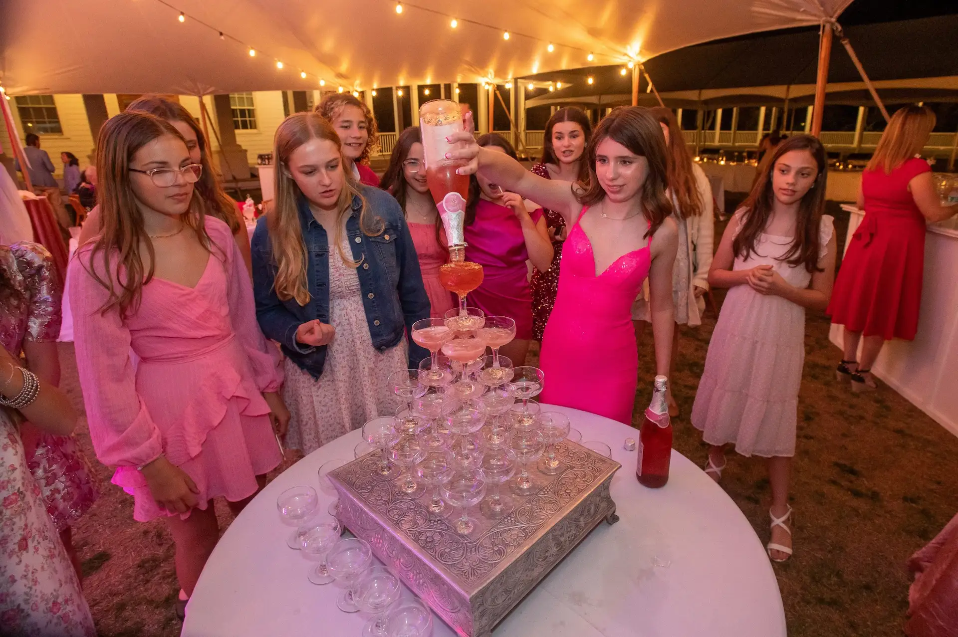 Guests watch as a girl pours sparkling drink into a champagne glass tower under lights.