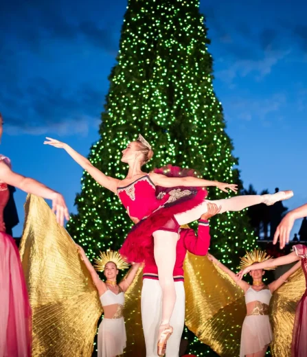 Outdoor holiday ballet performance with dancers in festive costumes in front of a large lit Christmas tree