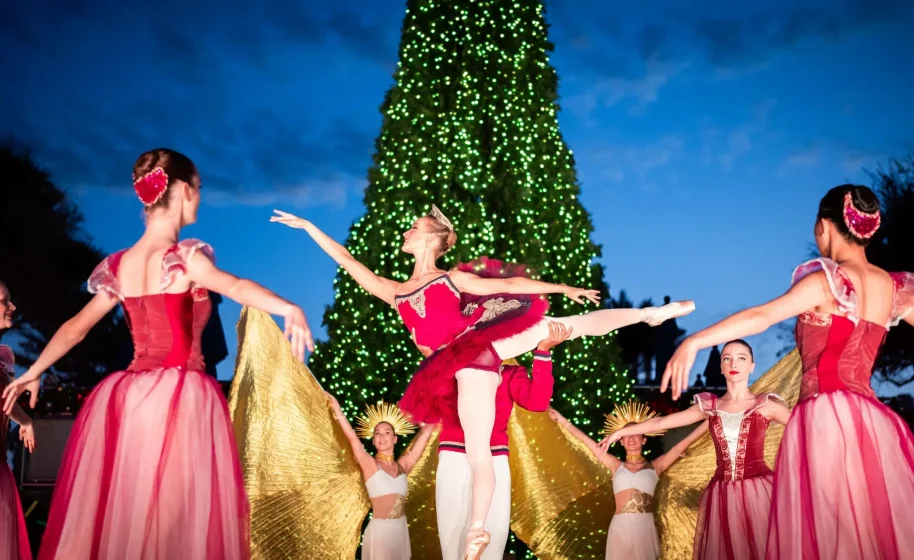 Outdoor holiday ballet performance with dancers in festive costumes in front of a large lit Christmas tree