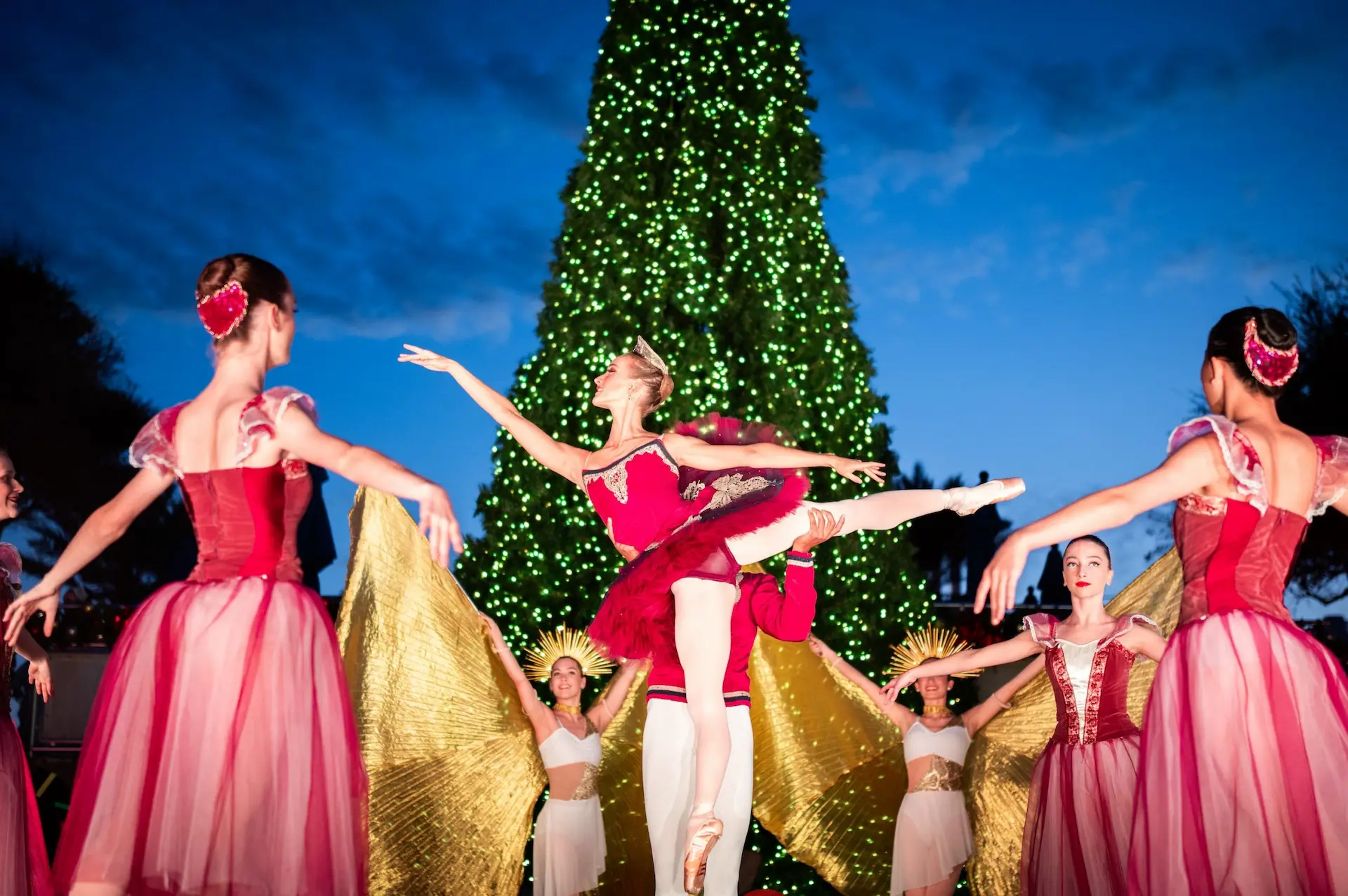 Outdoor holiday ballet performance with dancers in festive costumes in front of a large lit Christmas tree