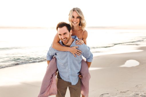 Smiling couple piggybacking on sunlit beach shoreline.