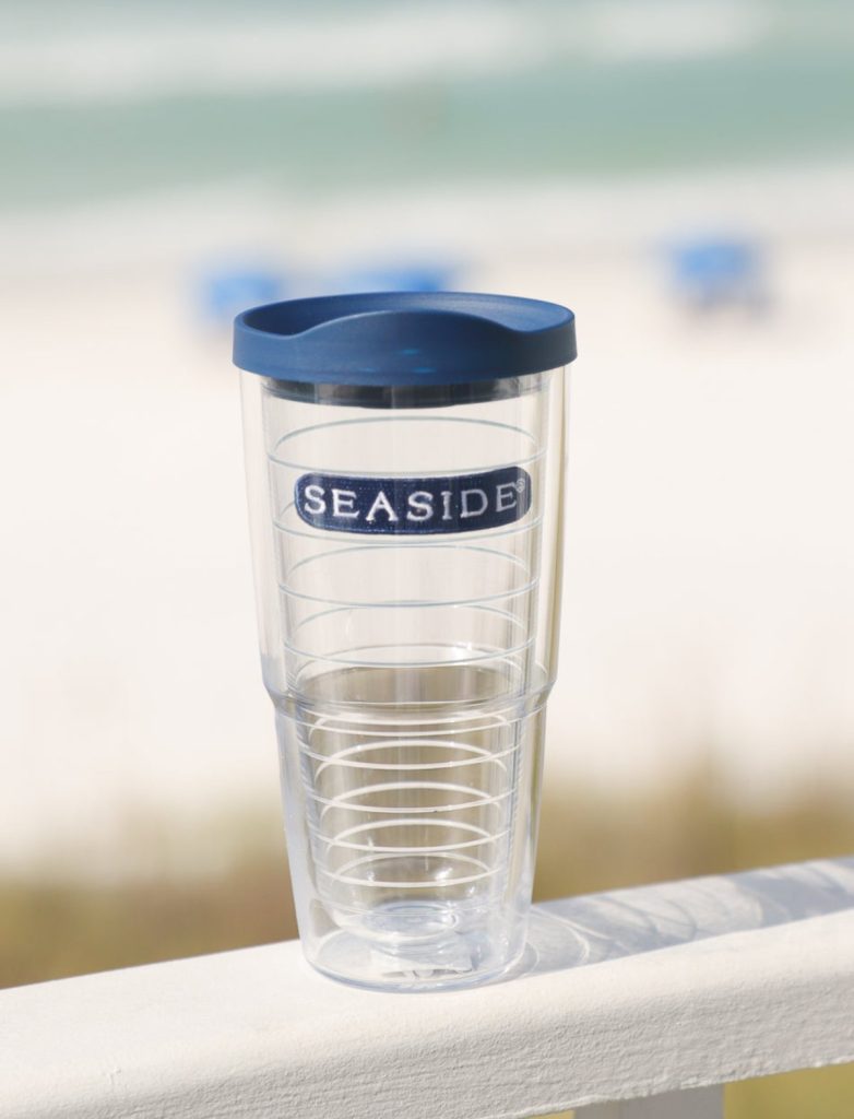 Clear double-walled tumbler with blue lid, labeled “Seaside,” rests on white railing with beach background.