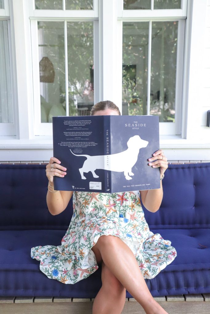 Woman wearing floral sundress sits on navy couch, reading book with dog silhouette illustration on cover.