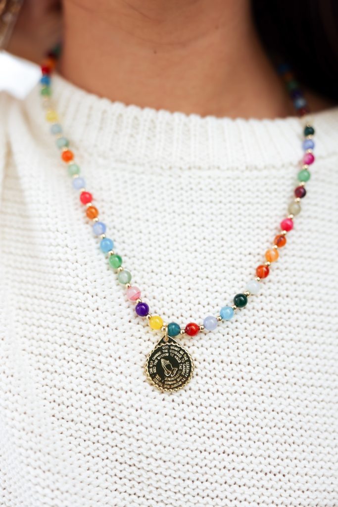 Close up of a colorful beaded necklace with round gold pendant, worn over a white knit sweater.