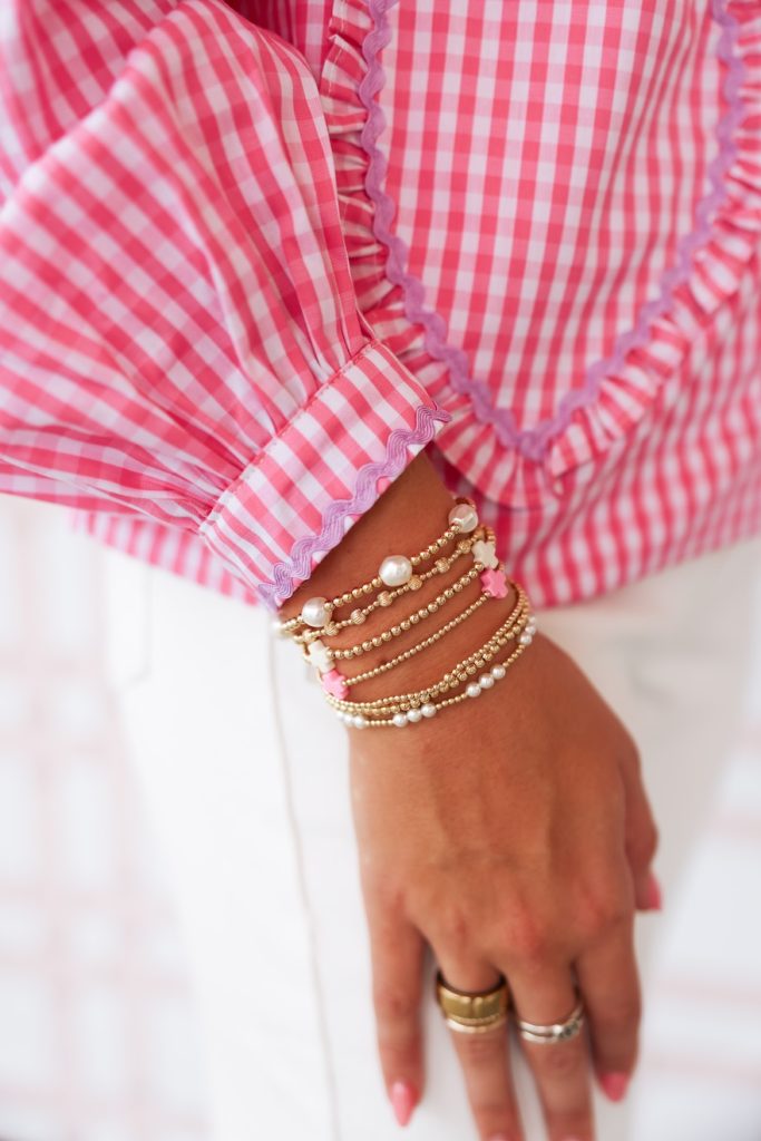 Woman’s hand and wrist adorned with gold and pearl bracelets, wearing a pink gingham shirt and white pants.