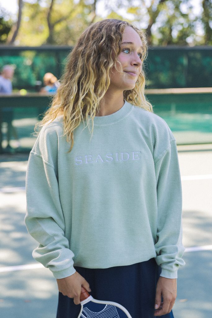 Woman holding a paddle, wearing light green ‘Seaside’ sweatshirt, standing on an outdoor tennis court.