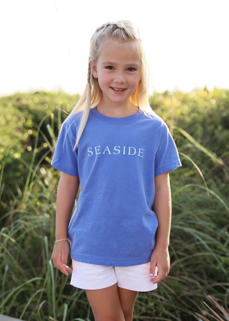 Smiling young girl wearing blue ‘Seaside’ T-shirt and white shorts standing outdoors in sunlight.