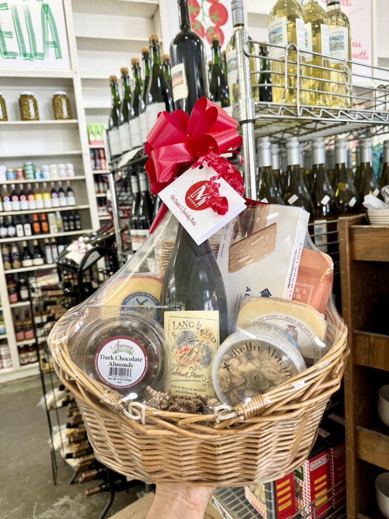 Handheld wicker gift basket with wine, cheese, crackers, chocolates, and corks, wrapped in cellophane with a red bow.