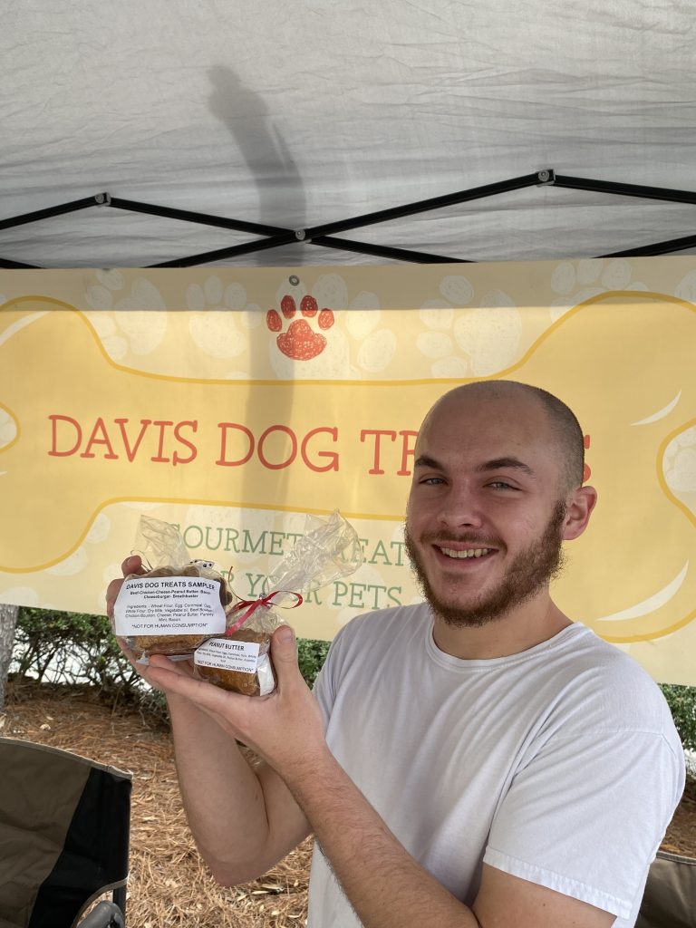 Vendor in white shirt holds packaged treats at Davis Dog Treats booth under yellow paw-print banner.