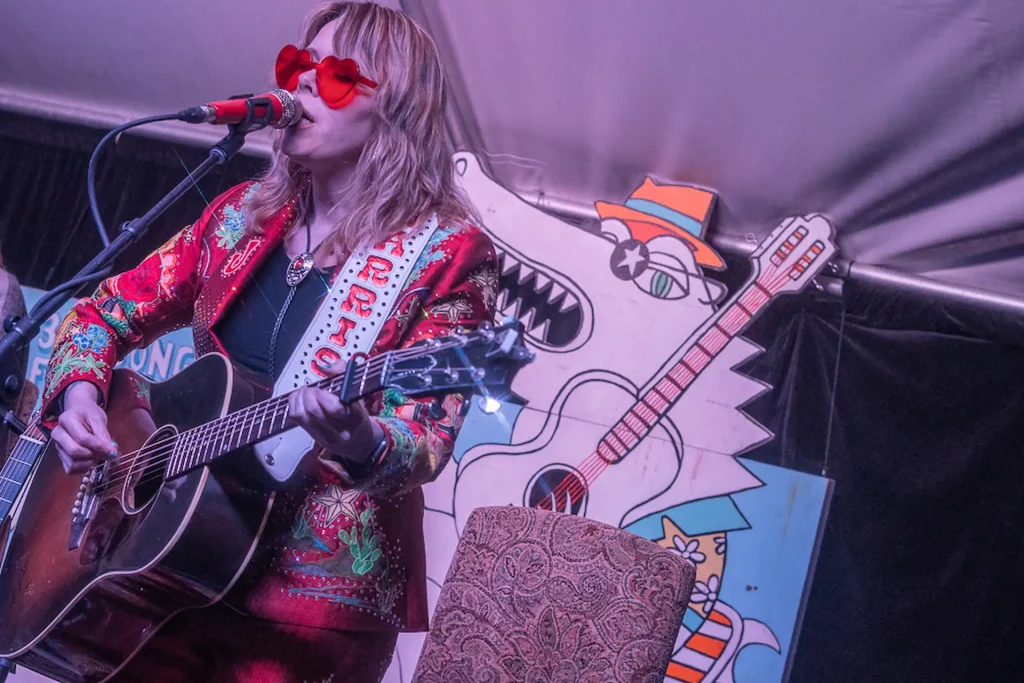 Singer in red rhinestone suit playing guitar onstage beside whimsical cartoon alligator backdrop.