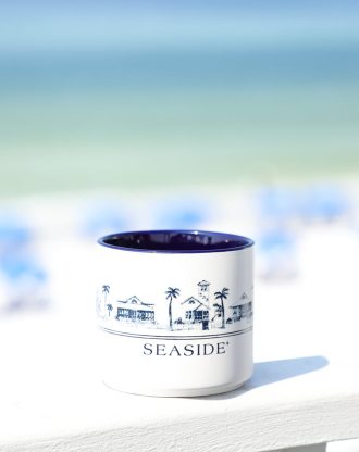 White mug with blue “Seaside” illustration of beach houses sits on ledge, blurred ocean and sky background.