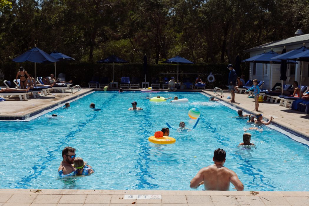 People swimming and relaxing in a sunny outdoor pool surrounded by lounge chairs and umbrellas.