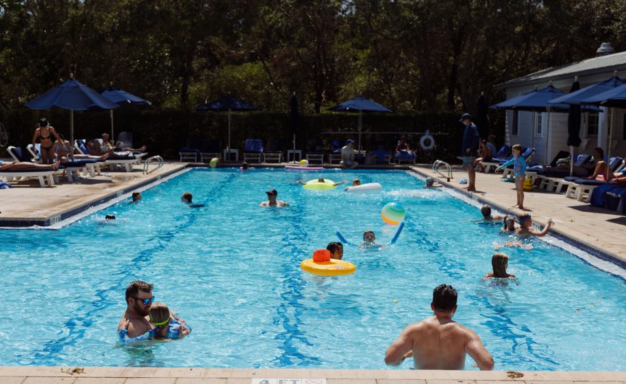 People swimming and relaxing in a sunny outdoor pool surrounded by lounge chairs and umbrellas.