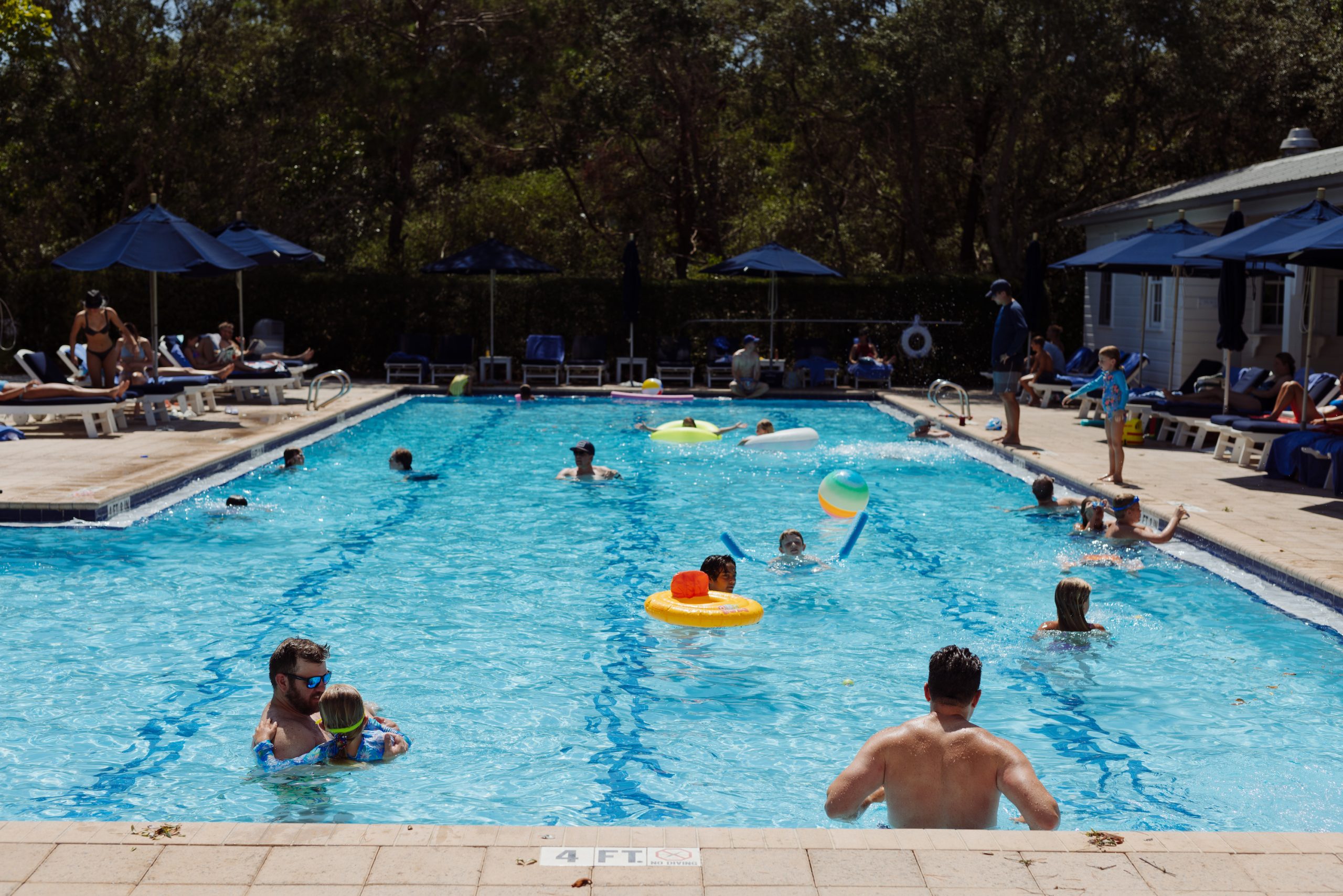 People swimming and relaxing in a sunny outdoor pool surrounded by lounge chairs and umbrellas.