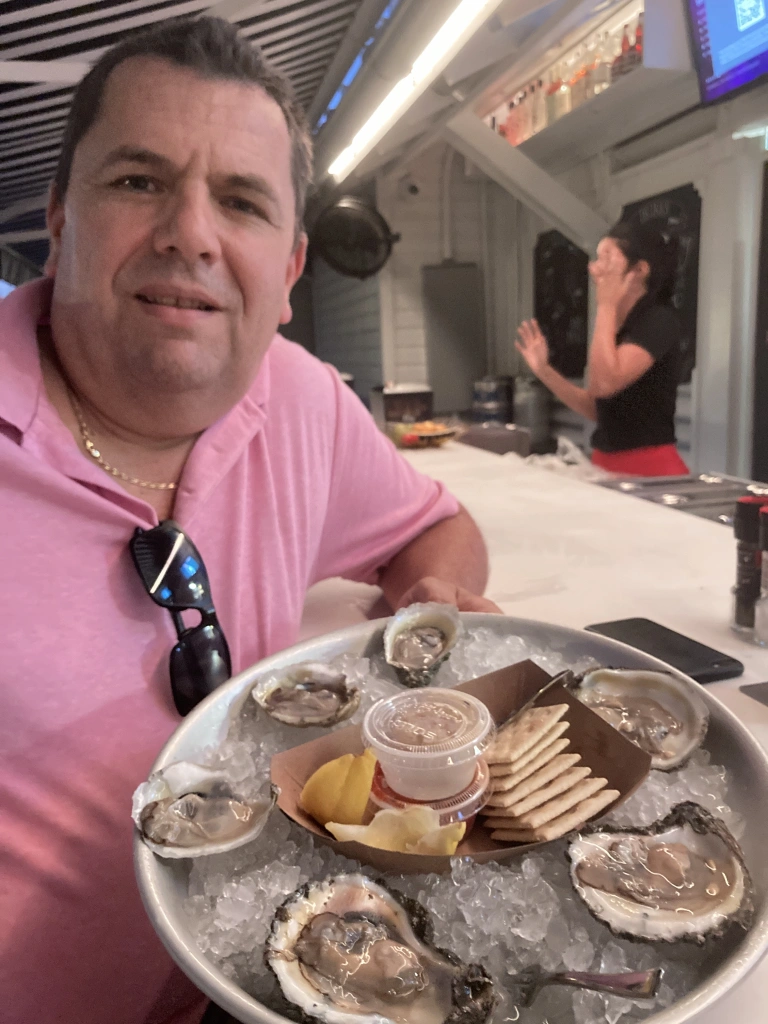 Man in a pink shirt holding a tray of oysters on ice with crackers and lemon.