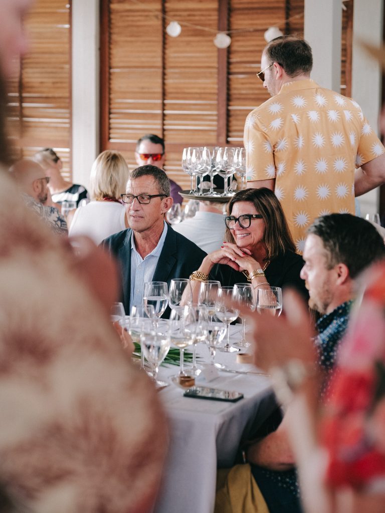 Guests seated at a dinner party while a server carries a tray of empty wine glasses.
