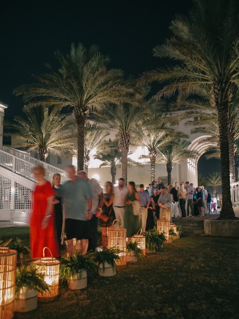 Evening crowd walking past lanterns and palm trees at an outdoor seaside event.