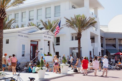 Groups of people gather outside the United States Post Office, with palm trees and bicycles on sunny day.