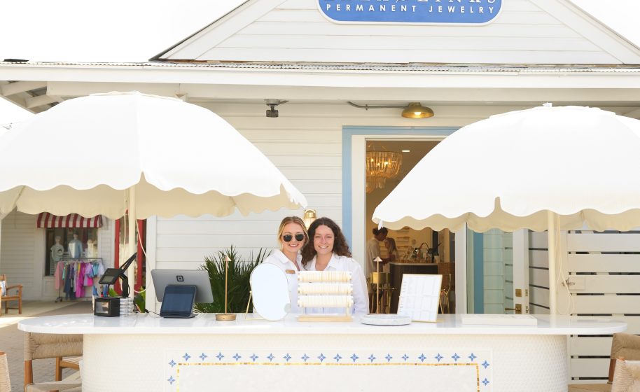 Two women standing at outdoor jewelry booth with umbrellas, Lala Links Permanent Jewelry shop sign visible above.