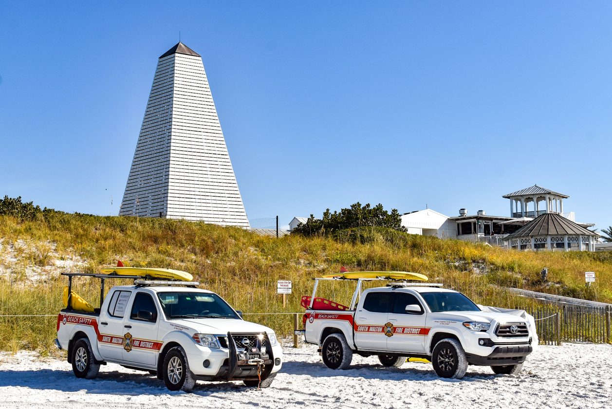 Two beach safety trucks parked on white sand below iconic seaside pavilion tower.