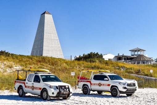 Two beach safety trucks parked on white sand below iconic seaside pavilion tower.