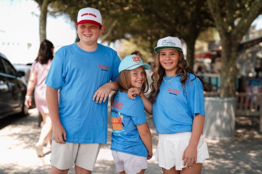 Three children smiling outdoors wearing matching blue T-shirts and baseball caps.