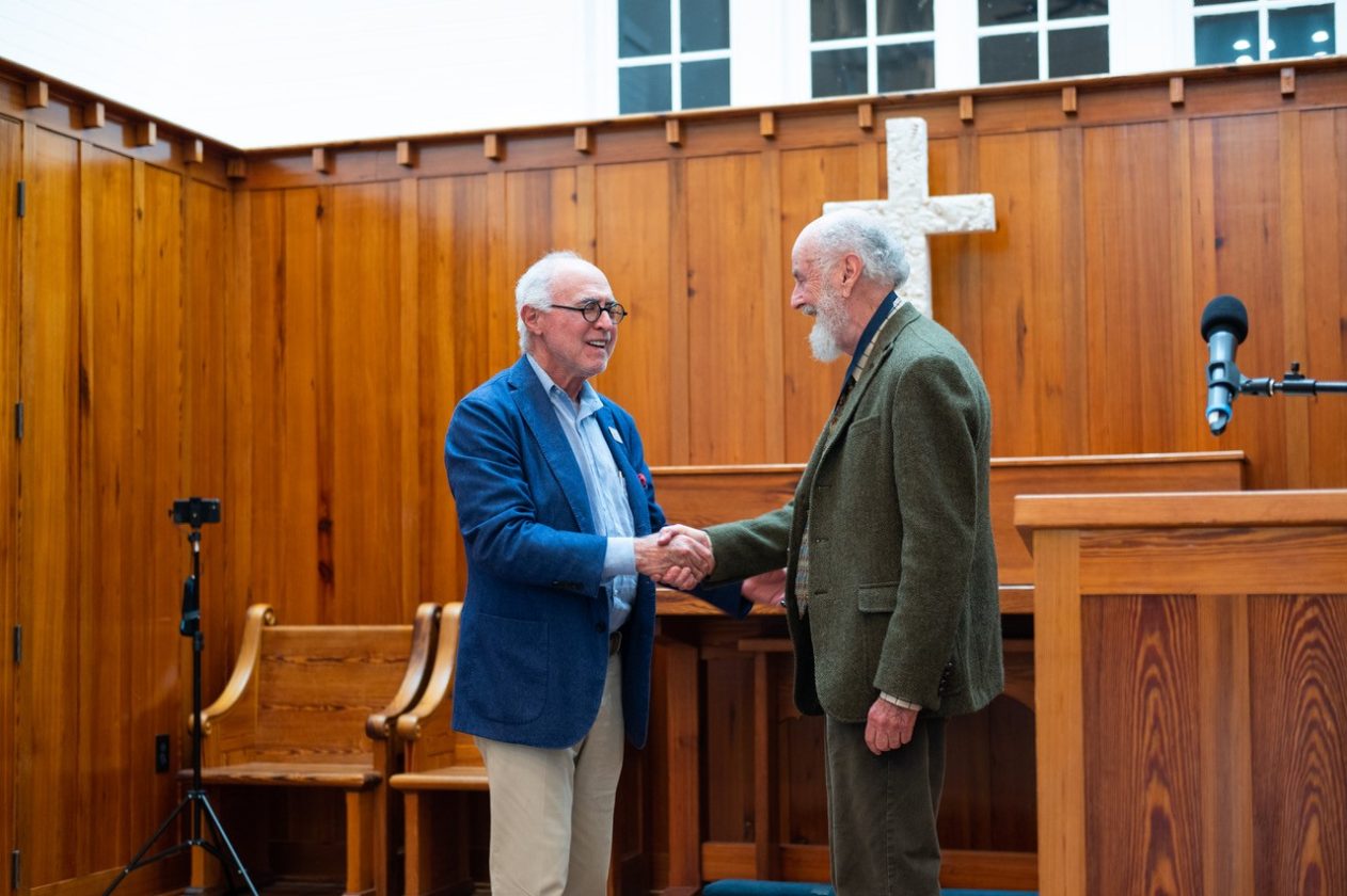Two elderly men smiling and shaking hands in a wooden chapel with a cross in the background.