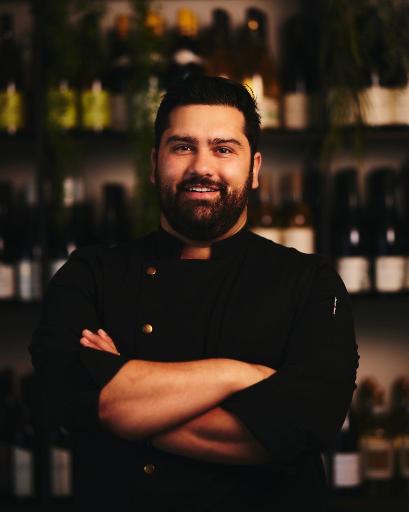 Smiling bearded chef with crossed arms standing before shelves of wine bottles.